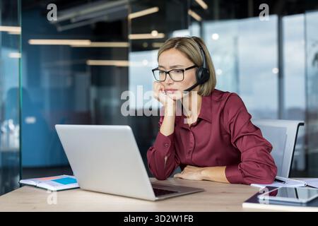 Kundendienstmitarbeiterin mit Headset und Brille, die müde und gelangweilt wirkt, während sie an ihrem Schreibtisch an einem Laptop arbeitet, was Stress, Ermüdung und Burnout hervorruft Stockfoto