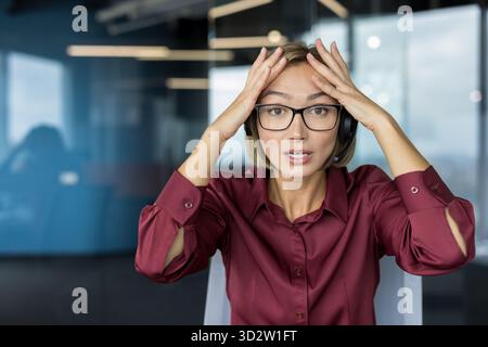 Gestresste Frau, die ein Headset und eine Brille trägt, den Kopf in den Händen hält, während sie in die Kamera blickt, und an einem Callcenter-Arbeitsplatz ein Problem oder einen Fehler erlebt Stockfoto