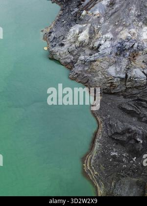 Aus der Vogelperspektive auf den atemberaubenden Kontrast zwischen dem türkisfarbenen See und dem dunklen, zerklüfteten vulkanischen Gelände, Mount Ijen, Ost-Java, Indonesien. Stockfoto