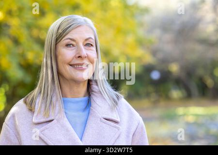 Ältere Frau mit grauen Haaren lächelt in einem Herbstpark selbstbewusst in die Kamera, trägt einen rosa Mantel und einen hellblauen Pullover, ruhig, gesund und genießt das goldene Laub der Natur Stockfoto