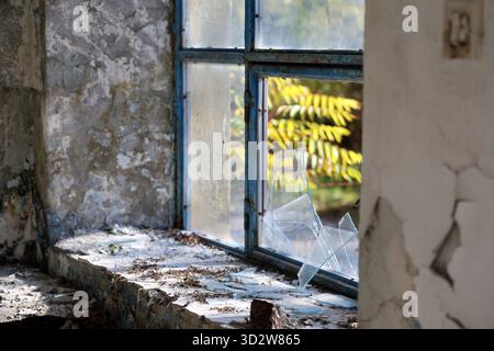 Der Blick von der Sonne durch ein blau gerahmtes, kaputtes Fenster zeigt abblätternde Gips und eine staubige Schwelle in einem verlassenen Innenraum, Schutt, Rost und bröckelnde Wände Stockfoto