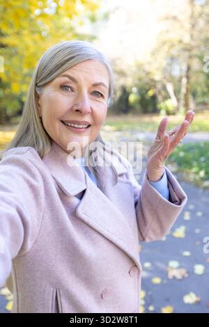 Die Seniorin filmt sich selbstbewusst bei einem Spaziergang in einem Herbstpark, hält ihren gesunden Lebensstil fest und teilt Momente mit moderner Technologie Stockfoto