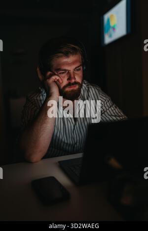 Mann mit Kopfhörern, der spät nachts am Laptop arbeitet, konzentriert und müde in einem dunklen Raum Stockfoto