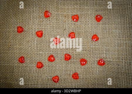 Glänzende rote Plastikherzen verstreut auf Sackleinen – romantischer Valentinstag Hintergrund Stockfoto