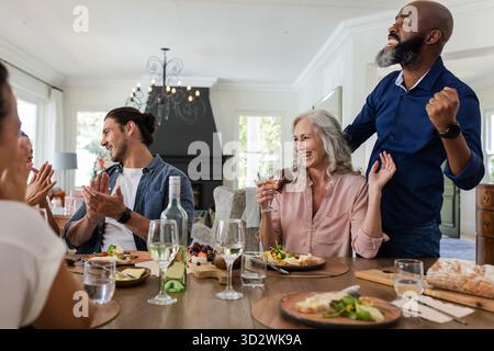 Freunde feiern am Esstisch, heben Gläser auf und klatschen im Haus. Feier, Dinnerparty, Toasten, Freundschaft, Treffen, Happiness || Model veröffentlicht Stockfoto