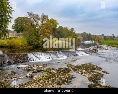 Linton Falls (ein Wehr) am River Wharfe in der Nähe des Dorfes Grassington, das das Kraftwerk Linton Hydro in der Ferne zeigt Stockfoto
