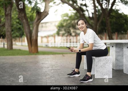 Eine Frau macht eine Pause auf einer Parkbank und lächelt, während sie ihr Handy überprüft. Stellt ein Gleichgewicht zwischen Fitness und Freizeit in einer natürlichen Umgebung dar Stockfoto