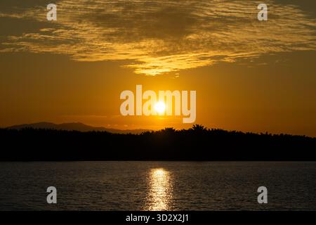 Ein atemberaubender goldener Sonnenuntergang über einem ruhigen Gewässer, mit einer dunklen Silhouette eines bewaldeten Horizonts und Bergen in der Ferne. Stockfoto