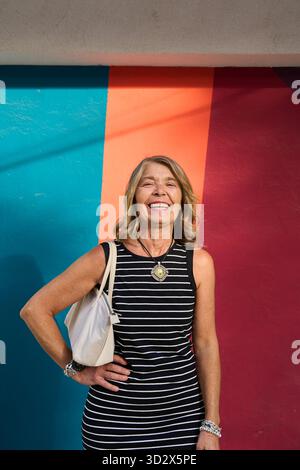 Glückliche ältere Frau, die steht, lacht und Freude zeigt, mit einer selbstbewussten Pose vor einer lebendigen, mehrfarbigen Wand Stockfoto