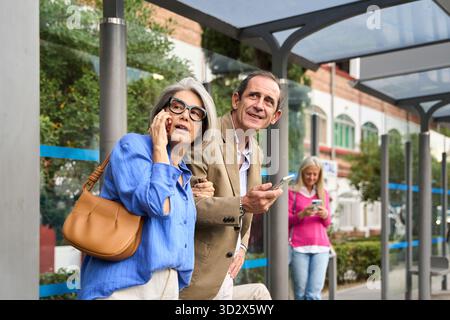 Seniorenpaar wartet an einer Bushaltestelle, eine Frau, die auf ihrem Telefon spricht, ein Mann, der sein Smartphone überprüft Stockfoto