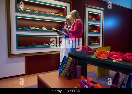 Zwei Seniorinnen wählen elegante Hausschuhe und Flats und genießen ein Shopping-Erlebnis in einer farbenfrohen Schuhboutique Stockfoto