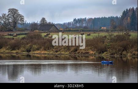 Eine Person in einem blauen Boot ruht auf einem breiten Fluss, mit einem Bauernhof und Herbstwald am entfernten Ufer Stockfoto
