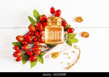 Eine Flasche mit natürlichem Hagebuttenkernöl liegt auf einem weißen Tablett zwischen einem Haufen reifer roter Früchte der Pflanze. Fruchtsamen in einem Löffel. Draufsicht Stockfoto