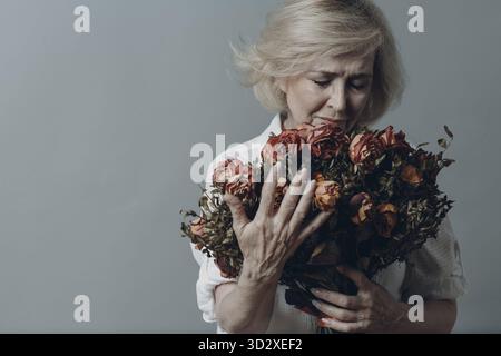 Traurige ältere Frau, die einen Strauß verwelkter Rosenblüten berührt. Grauhaarige reife Dame in Depression. Konzept des alten Alterns. Studioaufnahmen isoliert auf GRA Stockfoto
