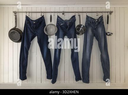 Drei Jeans hängen an einer Küchenstange zum Trocknen in einer kleinen Studentenwohnung. Minimalistisches, kreatives Leben, Mehrzweck. Weißer Wandhintergrund Stockfoto