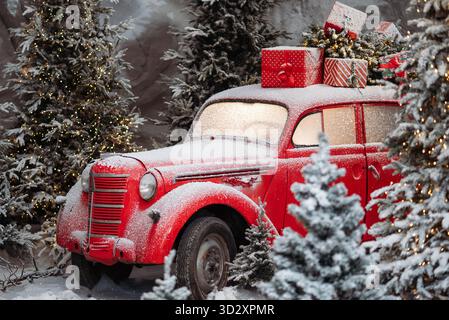 Roter Oldtimer mit Schnee, der Weihnachtsgeschenke und Baum in einem festlichen Winterwald mit Lichtern trägt Stockfoto