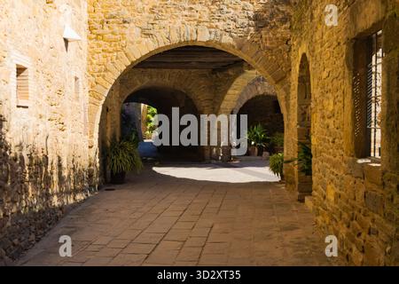 Steinbögen und Kopfsteinpflasterpfade in Monells, Katalonien, mit Topfpflanzen und warmem Sonnenlicht, die ein friedliches mittelalterliches Ambiente schaffen. Stockfoto