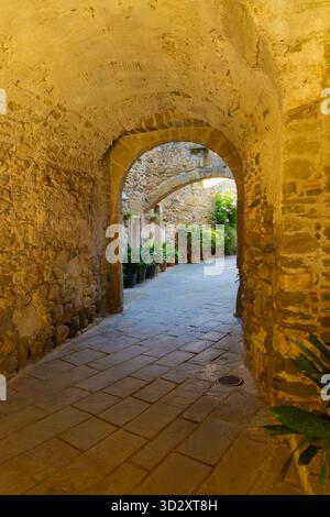 Steingasse mit Bögen in Monells, Katalonien, gesäumt von Topfpflanzen und in warmes Licht und historische Atmosphäre getaucht. Stockfoto