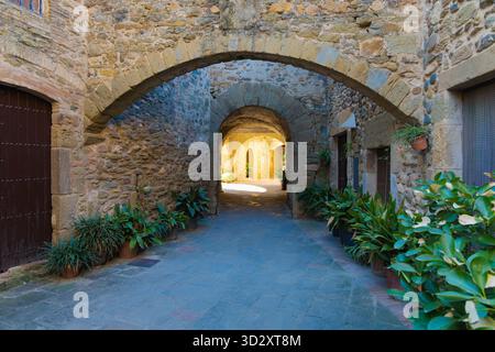 Eine Reihe von Steinbögen in Monells, Katalonien, säumen eine bezaubernde Gasse mit Topfpflanzen und sanftem goldenem Licht, die das mittelalterliche Ambiente unterstreichen. Stockfoto