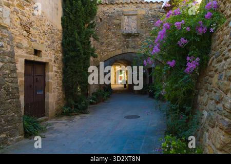 Mittelalterliche Steinallee in Monells, Katalonien, mit Bogengängen, Holztür, Topfpflanzen, und leuchtende rosa Blumen klettern an die Wand. Stockfoto