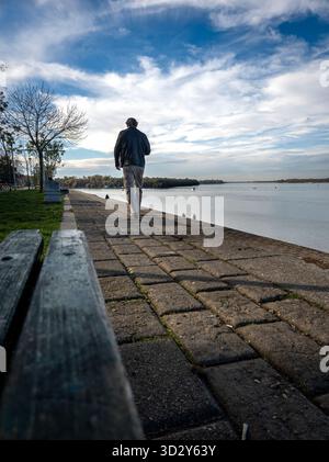 Ein Mann spaziert entlang der Uferpromenade an der Donau in Belgrad, Serbien, unter einem riesigen Himmel, der von weißen Wolken bedeckt ist, mit ruhigem Wasser, weit entfernt Stockfoto