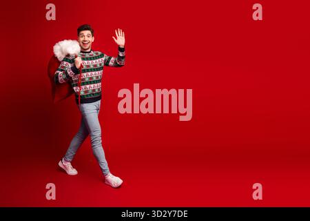 Lustiger junger Mann im festlichen Pullover trägt Santa-Tasche Wellen Hallo vor rotem Winterhintergrund für ein verspieltes Weihnachtsfoto Stockfoto