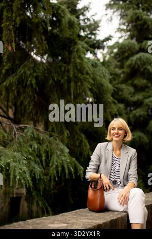 Eine lächelnde blonde Frau, die einen grauen Blazer, ein gestreiftes Hemd und eine weiße Hose trägt, sitzt draußen auf einem Steinvorsprung. Sie hat eine braune Tasche und genießt es Stockfoto