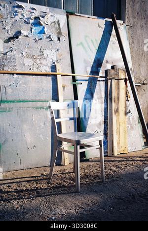 Alter Holzstuhl, silberfarben vor sonnendurchfluteten Metallpaneelen, in warmen analogen Tönen mit starker Textur und Kontrast. Stockfoto