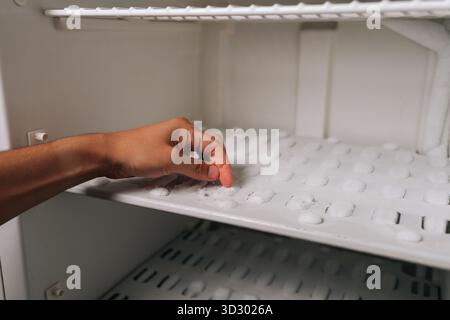 Die Fingerspitzen drücken gegen die dicke Frostschicht im Gefrierfach, wodurch starke Eisansammlungen sichtbar werden, die dringend zum Abtauen erforderlich sind. Stockfoto