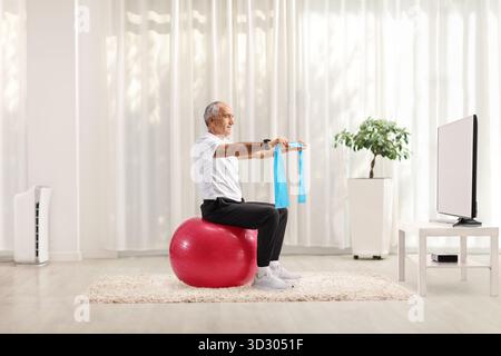 Reifer Mann, der mit einem Widerstandsband trainiert und zu Hause auf einem Fitnessball vor dem fernseher sitzt Stockfoto