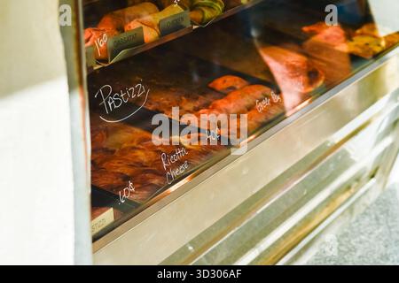 Ein Blick in eine hell beleuchtete Glasvitrine einer traditionellen maltesischen Bäckerei mit verschiedenen Arten frisch gebackener, herzhafter Pastizzi. Handgeschriebene Etiketten weisen auf Füllungen wie Ricotta-Käse hin. Stockfoto