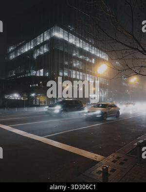 Verkehr in der Stadt bei Nacht. Straßen mit dichtem Nebel und hellen Lichtern vorbeifahrender Autos. Modernes Business Center im Hintergrund. Stockfoto