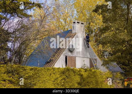Ein Arbeiter klettert auf einer Leiter auf einem Haus mit Steildach, das gerade renoviert wird und von Herbstbäumen und Hecken umgeben ist. Das Dach ist teilweise mit neuen Fliesen bedeckt Stockfoto