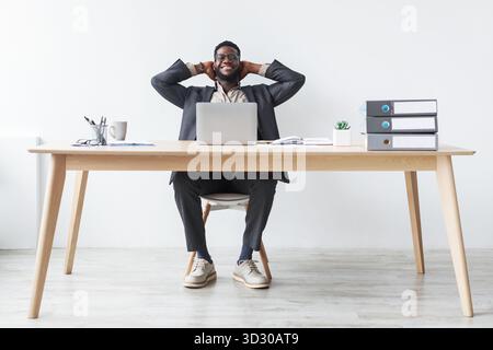Entspannter junger schwarzer Geschäftsmann macht eine Pause am Schreibtisch, während er ein Notebook benutzt Stockfoto