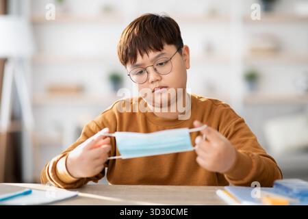 Der gelangweilte asiatische Teenager kämpft mit der Schule, während er zu Hause eine schützende Gesichtsmaske hält Stockfoto