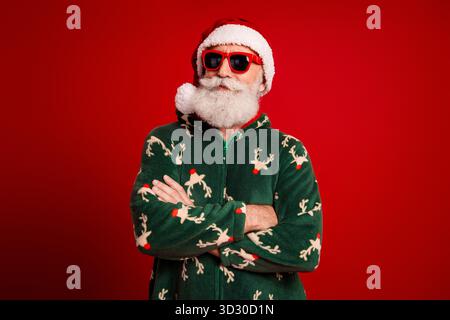 Weihnachtsmann in grünem weihnachtspullover mit roter Sonnenbrille steht selbstbewusst vor einem kräftigen roten Hintergrund Stockfoto