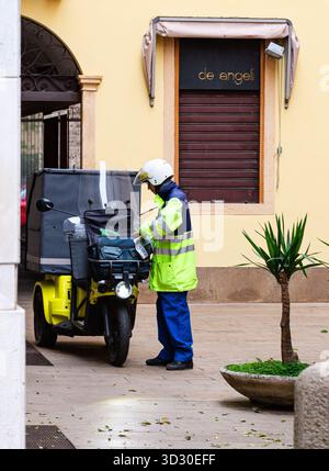 Padua, Italien - 14. April 2025: Lieferarbeiter in Sicherheitskleidung organisiert Pakete auf einem gelben Roller in einer pulsierenden städtischen Umgebung mit Pflanzen Stockfoto