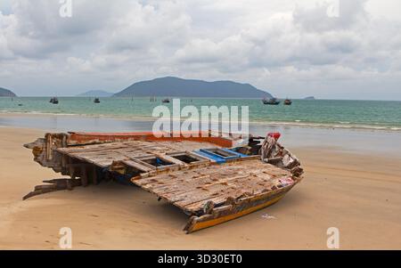 Verlassenes hölzernes Schiffswrack an einem Sandstrand mit Fischerbooten und Inseln im Hintergrund unter bewölktem Himmel. Stockfoto