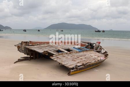 Verlassenes hölzernes Fischerboot-Wrack an einem breiten Sandstrand in Con Dao, Vietnam, mit Fischerbooten und Inseln am Horizont. Stockfoto