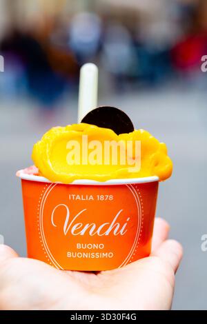 Padova, Italien - 14. April 2025: Hand präsentiert eine bunte Eisbecher aus Venchi mit Mangogeschmack und Schokoladenscheibe, ideal für warme Tage Stockfoto