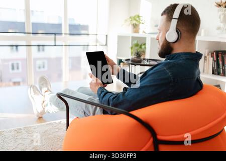 Komfortabler Mann, der sich mit einem Tablet im modernen Wohnzimmer entspannt und Kopfhörer trägt Stockfoto