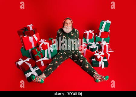 Der fröhliche Weihnachtsmann im gemütlichen Pyjama sitzt zwischen Geschenkboxen auf rotem Hintergrund und ist bereit für Weihnachtsfeiern, Weihnachtseinkäufe und Familienmomente Stockfoto