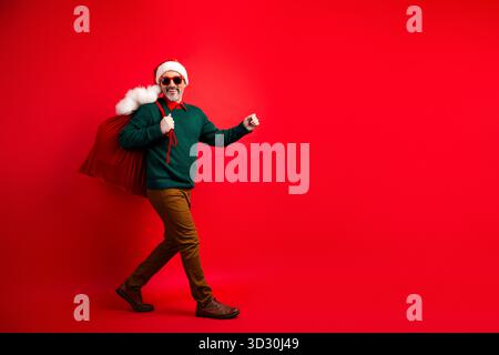 Der lustige Weihnachtsmann trägt eine große rote Tasche mit einem grünen Pullover und einer Sonnenbrille vor einem knallroten Hintergrund Stockfoto
