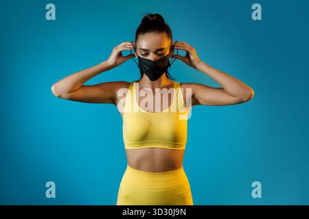 Sportliche Frau, die eine Gesichtsmaske aufsetzt, während sie gelbe Fitwear vor blauem Hintergrund trägt Stockfoto