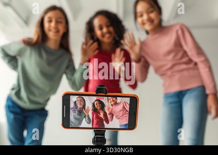Drei multikulturelle Mädchen kreieren drinnen einen lustigen Video-Blog, während sie der Kamera zuwinken Stockfoto