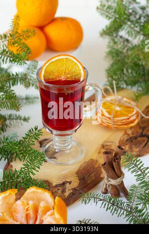 Ein warmes Gewürzgetränk in einem klaren Glas sitzt auf einer Holzoberfläche. Getrocknete Orangenscheiben und frische Orangen umgeben das Glas und verleihen dem Glas Farbe und ein festliches Ambiente Stockfoto