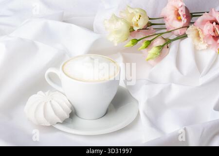 Herzerwärmende Szene mit einer zarten Kaffeetasse mit aufschäumender Milch und einem Meringue-Leckerbissen, ergänzt durch wunderschöne Blumen auf weißem Stoff, cr Stockfoto