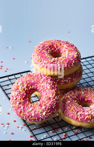 Vier rosafarbene Donuts mit bunten Streuseln sind auf einem Kühlregal aus Metall gestapelt. Bunte Streusel streuen sich herum und sorgen so für Spaß Stockfoto