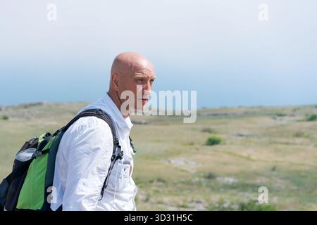 Ein Mann mit einem Glatzkopf trägt ein weißes Hemd und einen Rucksack. Er steht auf einem Feld Stockfoto