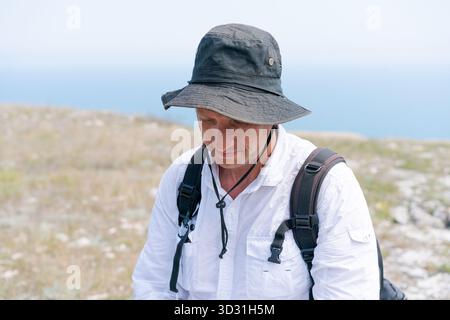 Ein Mann mit schwarzem Hut und weißem Hemd sitzt auf einem Hügel. Er hat einen Rucksack auf dem Rücken Stockfoto